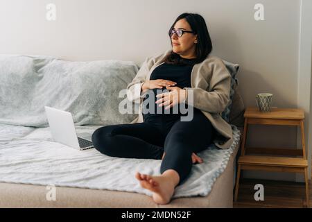 Pregnant woman freelancer sitting at home on the couch working on a laptop. Stock Photo