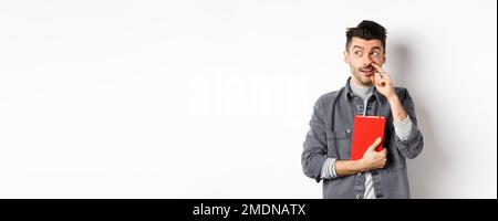 Guy picking nose and look aside at logo, holding red book or planner in hand, standing against white background Stock Photo