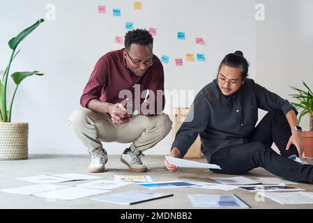 Anythings possible with a proper plan. Full length shot of two young businessmen sitting down on the floor and going over paperwork together at work. Stock Photo