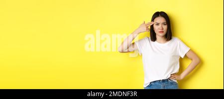 Woman showing kill me please gesture, shooting herself in head with finger gun from boredom, standing upset over yellow background Stock Photo