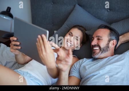 Sweet memories of their very first date. High angle shot of an affectionate couple using a digital tablet while lying in bed together in the morning. Stock Photo