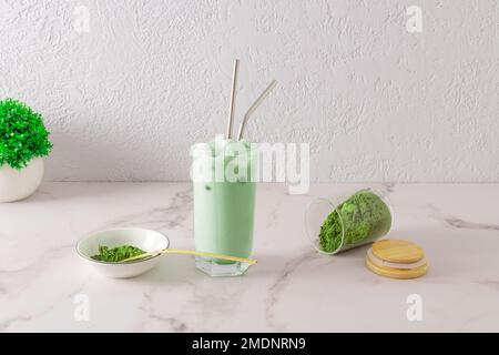 A tall glass of organic green tea matcha latte with eco-friendly metal straw stands on a marble table. scattered tea powder from a jar Stock Photo