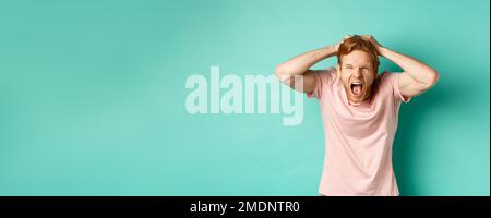 Angry redhead man screaming and ripping hair out of head, yelling and losing temper, looking insane from rage, standing over mint background Stock Photo