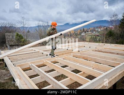 Man worker building wooden frame house on pile foundation. Carpenter carrying wooden board. Carpentry concept. Stock Photo