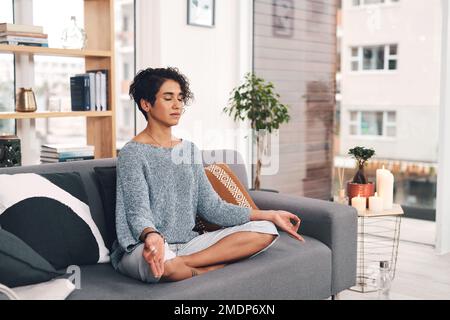 The journey outwards is inwards. Full length shot of an attractive young woman sitting and meditating on her sofa in the living room at home. Stock Photo