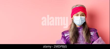 Covid-19, virus and social distancing concept. Sassy asian senior woman in medical mask and glitter dress winking, wearing party outfit, pink Stock Photo