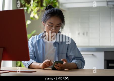 Young Asian female freelancer scrolling her phone, getting distracted while freelancing at home Stock Photo