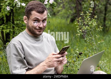 Man with cunning eyes wearing glasses uses his cell phone and laptop while sitting outdoors on green grass looking at camera. Stock Photo