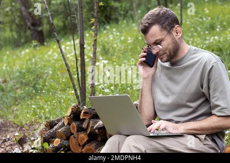 Young businessman working outdoors calling cell phone and using his laptop. Male person using digital devices in nature. Stock Photo