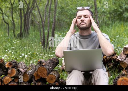 Man with closed eyes holding head in hands while working on laptop outdoors sitting on firewood pile. Stock Photo