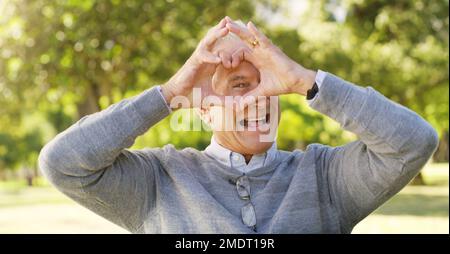 Hes looking at you through the eyes of love. Cropped portrait of a happy senior man showing the love symbol at the park. Stock Photo