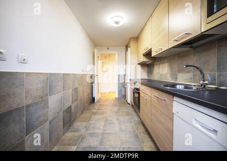 Elongated kitchen with wooden furniture with a black top, gray tiles, a circular lamp on the ceiling and white wooden access doors Stock Photo
