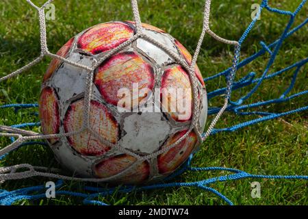 old soccer ball in the net on the background of grass soccer field. Summer sunny day. Stock Photo