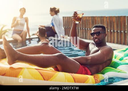 The coolest guy at the party. Portrait of handsome young man raising up his glass for a toast while relaxing in a pool outdoors with friends. Stock Photo