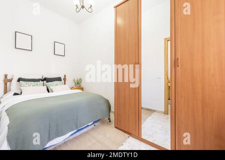 Room with a double bed with a green blanket, lots of cushions, a three-section wardrobe with a central mirror door and a wooden headboard Stock Photo