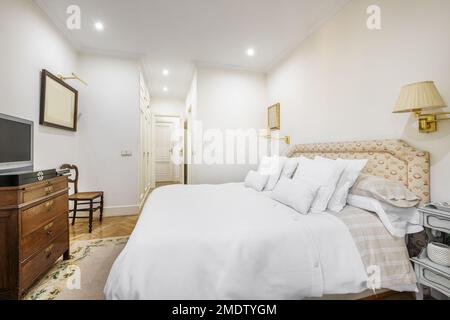 A room with a double bed decorated in a classic style with a headboard upholstered in fabrics and many cushions of different sizes with white covers Stock Photo