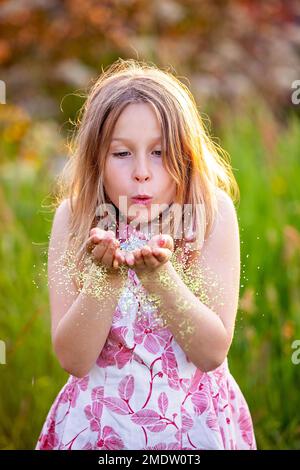 Young girl, child, stunning blond kid with long blonde hair locks, blowing a magic golden powder to the camera. Fairy tale, witch, imaginative photo. Stock Photo