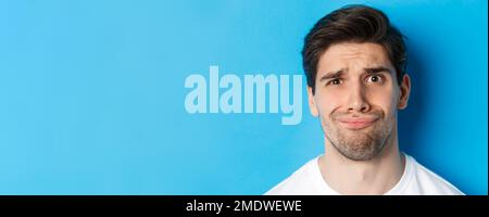 Head shot of skeptical and disappointed man, grimacing complicated, standing against blue background Stock Photo
