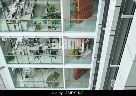 Part of modern multi-storey building with rows of desks, armchairs and green plants in openspace offices behind large windows Stock Photo