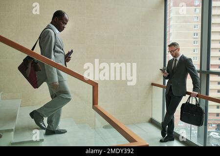 Mature African American businessman walking downstairs while young male entrepreneur using smartphone inside modern business center Stock Photo