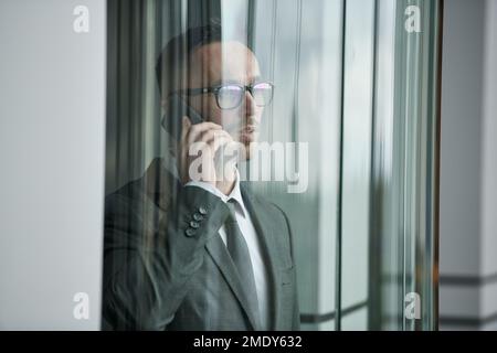 Young businessman in formalwear and eyeglasses speaking on mobile phone in transparent glass elevator inside modern office center Stock Photo