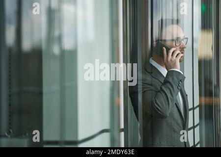 Young confident male entrepreneur in elegant dark grey suit and eyeglasses talking on mobile phone while moving in elevator Stock Photo