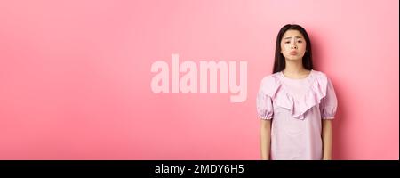 Sad whining asian girl pouting and frowning, looking upset about something unfair, complaining, standing in dress against pink background Stock Photo