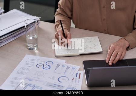 Close-up of hands of female entrepreneur analyzing graphic data and making notes in notebook while working over new project Stock Photo
