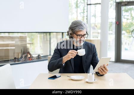 Bearded mature economist or banker in suit and eyeglasses analyzing online financial data in tablet by cup of coffee in modern restaurant Stock Photo