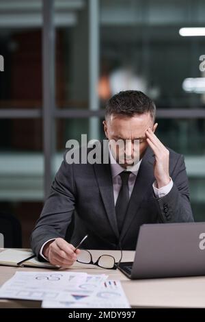 Overworked and tired businessman in formalwear sitting by desk, bending over laptop and touching temple while trying to concentrate Stock Photo