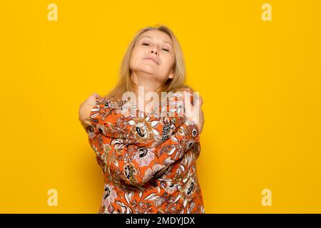 Close-up photo of a charming woman wearing a dress while smiling and hugging herself, isolated over yellow background. Stock Photo