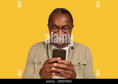 Cheerful african american middle aged male in glasses typing on smartphone, surfing in internet Stock Photo