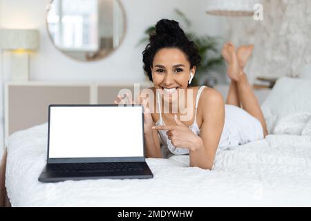 Smiling arab millennial female woke up, lies on bed in wireless headphones, points finger at laptop Stock Photo
