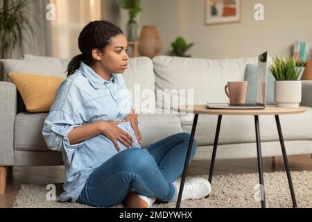 Young black pregnant woman video calling via laptop and touching her belly, communicationg with doctor online Stock Photo