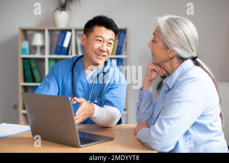Happy glad handsome middle aged korean doctor shows laptop to old female caucasian patient in clinic Stock Photo