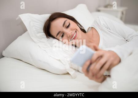 Happy young lady holding smartphone while lying in bed Stock Photo