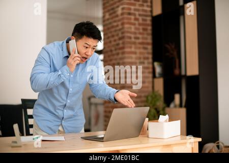 Stressed mature asian businessman talking on cellphone and looking at laptop, working at home office, free space Stock Photo
