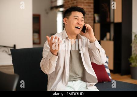 Happy mature asian man talking on cellphone and gesturing, having pleasant conversation while sitting on sofa at home Stock Photo
