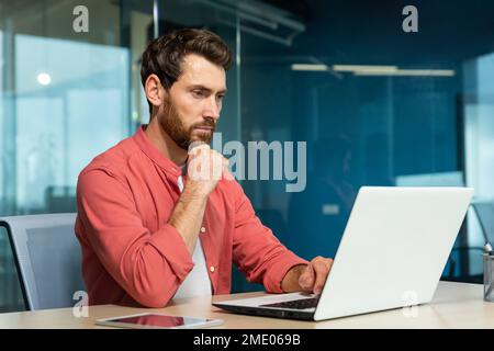 Serious thinking businessman close up, man in red casual shirt working inside modern office, mature boss working with laptop at workplace while sitting Stock Photo