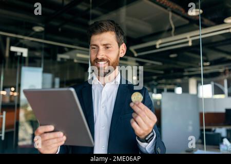 Businessman looking at tablet and holding bitcoin coin, analyzing financial data at workplace in office interior Stock Photo