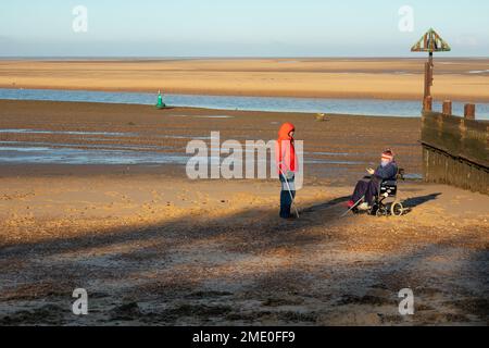 Lady in wheelchair and person with crutch walking aid on beach during winter at Wells on sea North Norfolk East Anglia, England Stock Photo