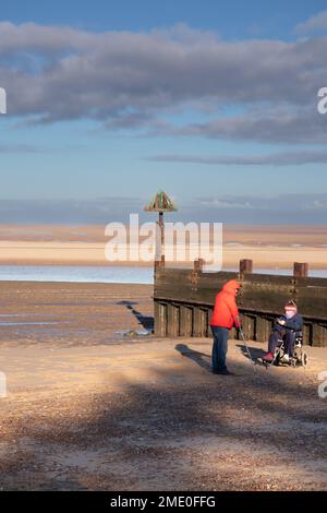 Lady in wheelchair and person with crutch walking aid on beach during winter at Wells on sea North Norfolk East Anglia, England Stock Photo
