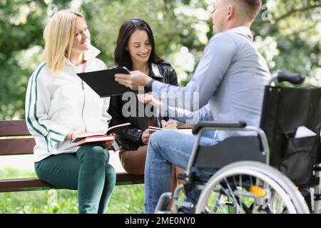 Two female students and one male student study together in park, man is sitting in wheelchair. Stock Photo
