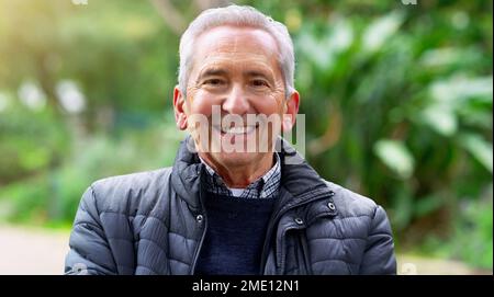Always a optimistic one. a cheerful elderly man seated on a bench outside in a park during the day. Stock Photo