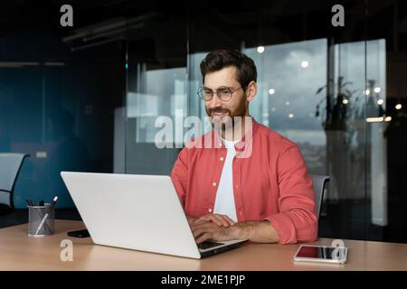 Successful smiling man working inside office with laptop, businessman in red shirt smiling and typing on keyboard in glasses, programmer working software for program. Stock Photo
