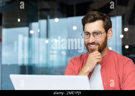 Successful smiling man working inside office with laptop, businessman in red shirt smiling and typing on keyboard in glasses, programmer working software for program. Stock Photo