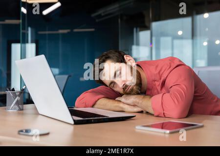 Tired businessman in casual red shirt sleeps at workplace, man on desk fell asleep during working hours inside office with laptop. Stock Photo