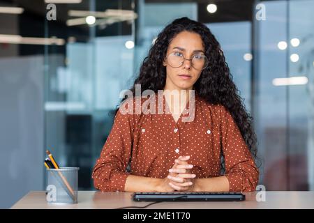 Portrait of a young Hispanic woman teacher. She sits seriously at the tablein the office , folded her hands on the table, looks into the camera. Stock Photo