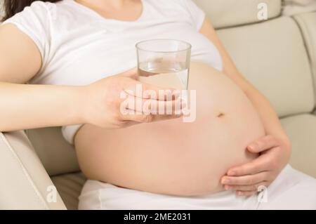 An pregnant woman sitting in the live room thinking and holding a glass of water Stock Photo