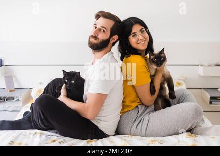 Side view of happy young couple in domestic clothes resting on bed together while hugging cats and smiling at weekend Stock Photo